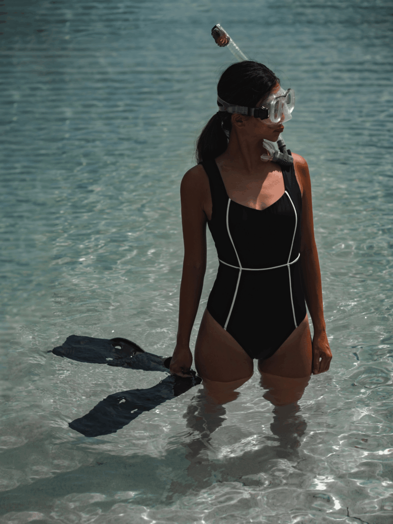 Woman with a snorkel mask wading in clear water holding fins in the black Alma one-piece