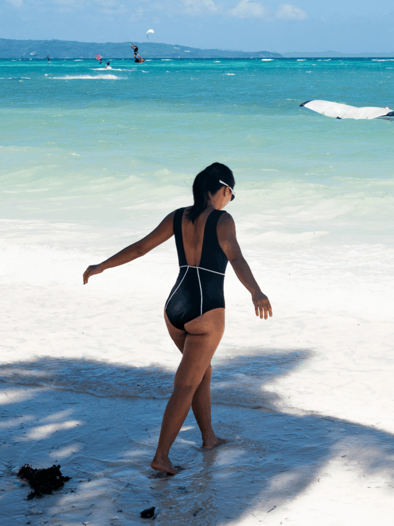 Woman walking on a white sand beach toward the sea in the black Alma one-piece
