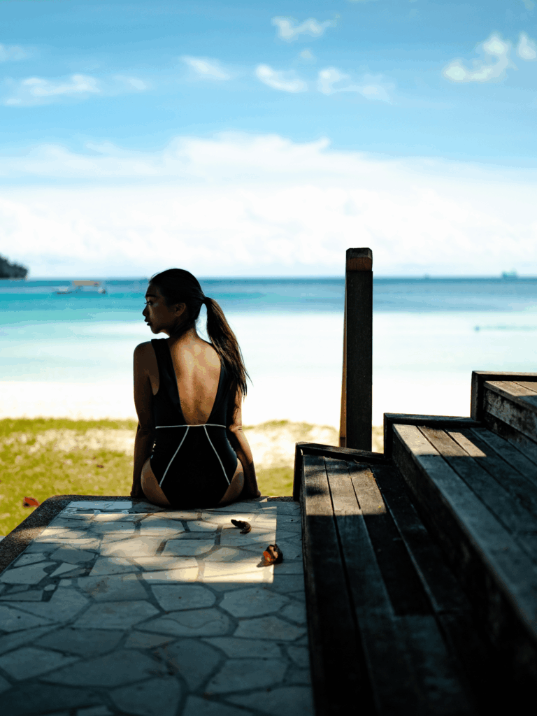Woman sitting on a wooden deck facing the sea in the black Alma one-piece