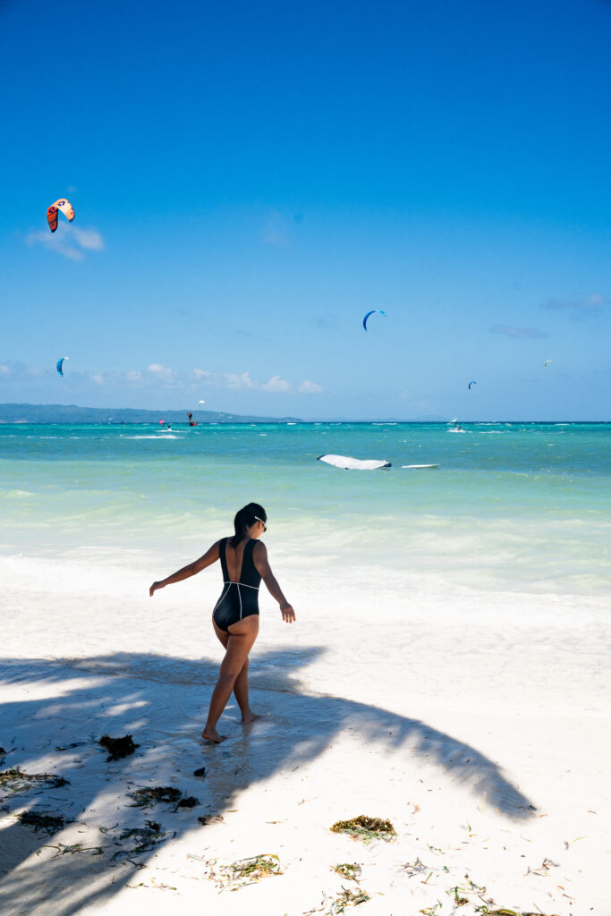 Woman walking on a white sand beach in the black Alma one-piece with white piping