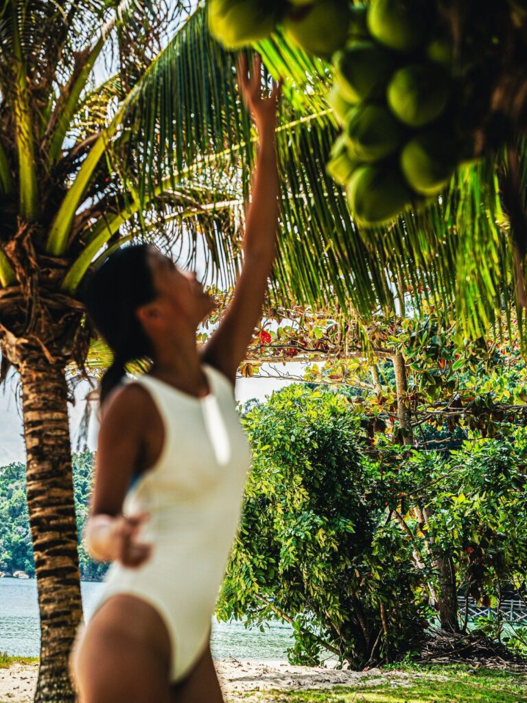 Woman reaching for coconuts under a palm tree in the cream white Pona one-piece