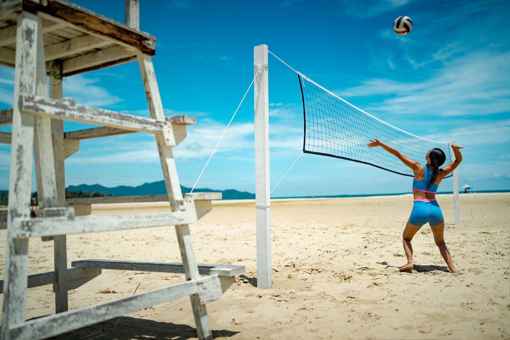 Woman serving a volleyball on the beach in the azure blue Tenacy two-piece