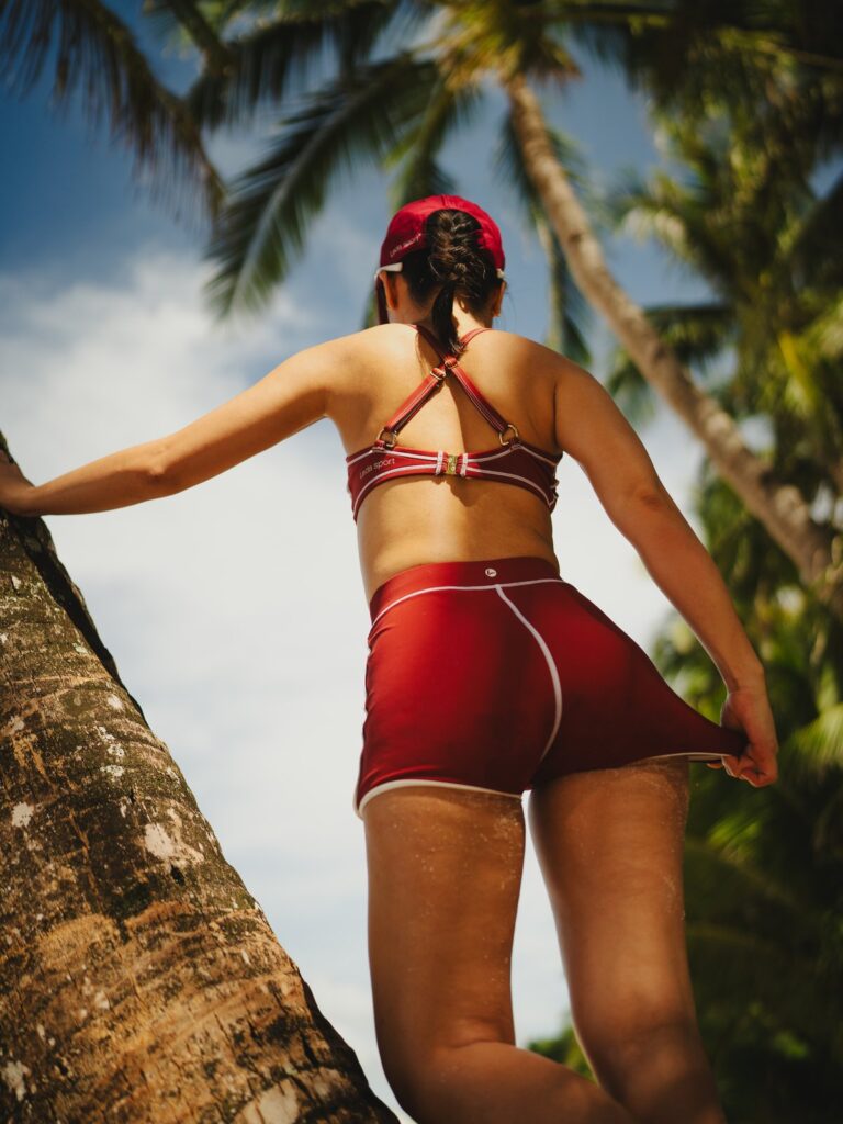 Woman leaning on a palm tree on the beach in the Cala boyshort, seen from the back