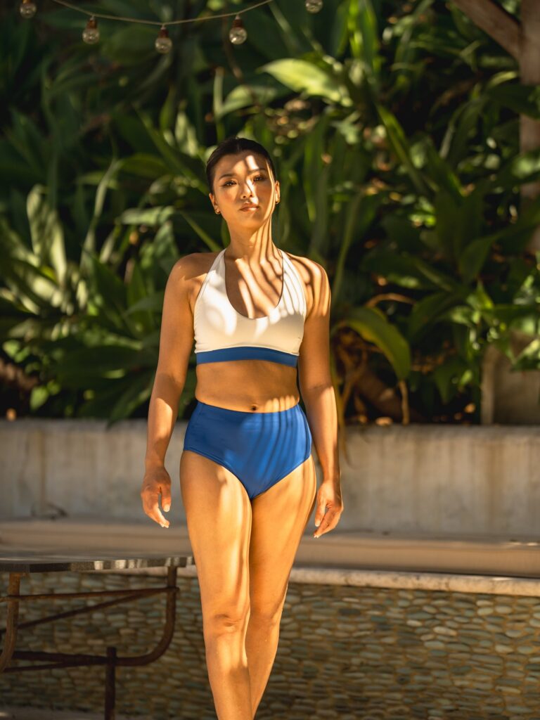 Woman walking beside a pool in the cream white Cala swim top with a royal blue band