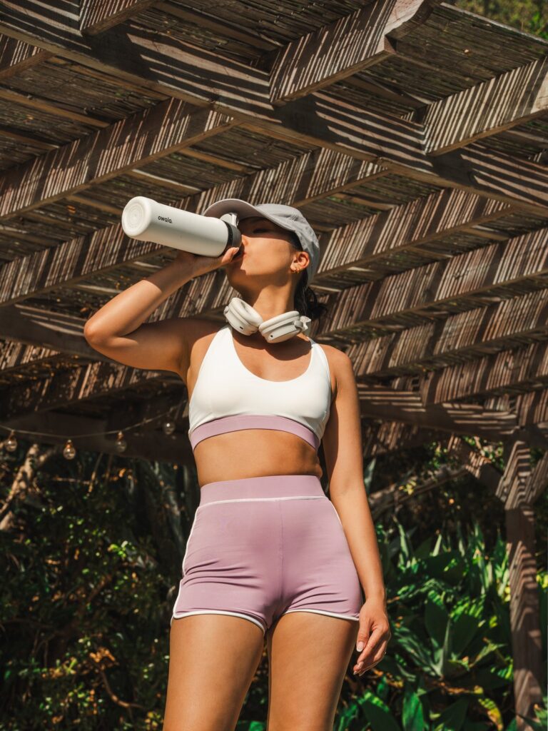 Woman drinking from a water bottle under a pergola in the cream white Cala swim top